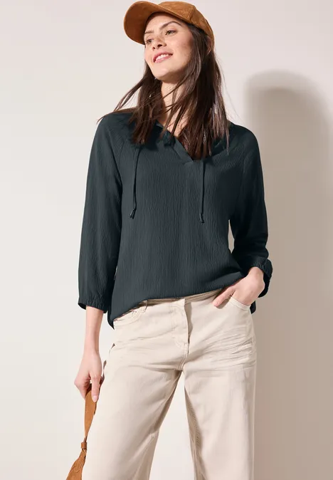 Frau mit brauner Kappe, schwarzer Bluse und heller Hose, lächelt.